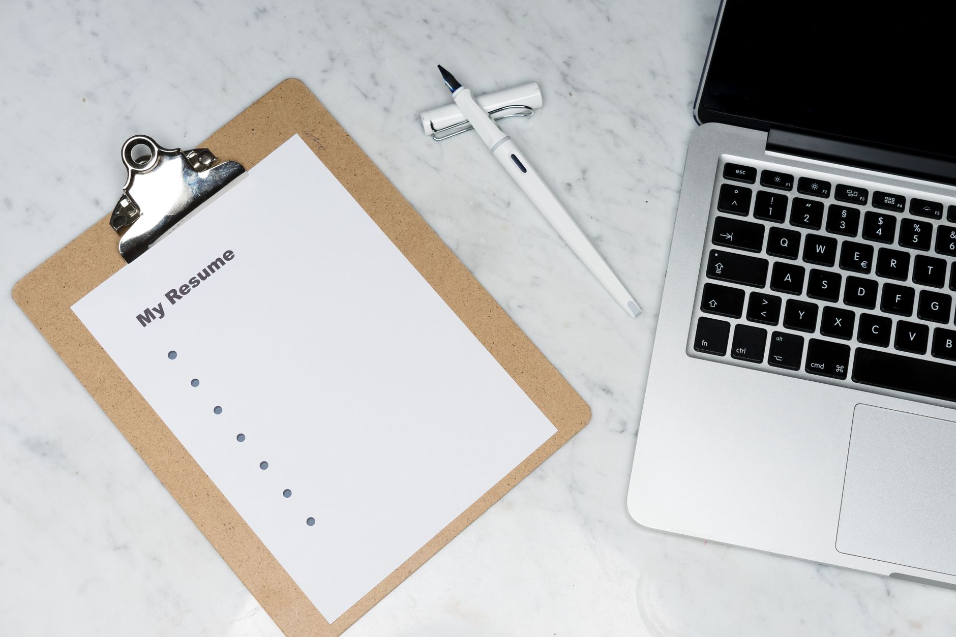 A clipboard, pen, and laptop on a marble desk representing resume writing